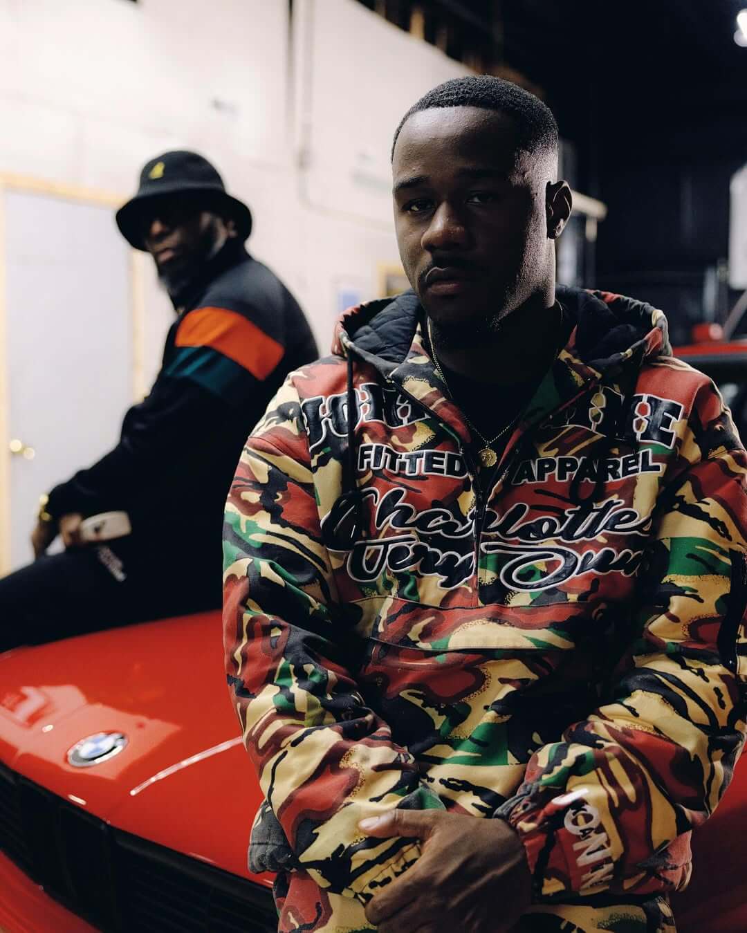 Reuben Vincent in camouflage leaning against a red BMW with 9th Wonder, his mentor in the background for their single "Gotta Get It."