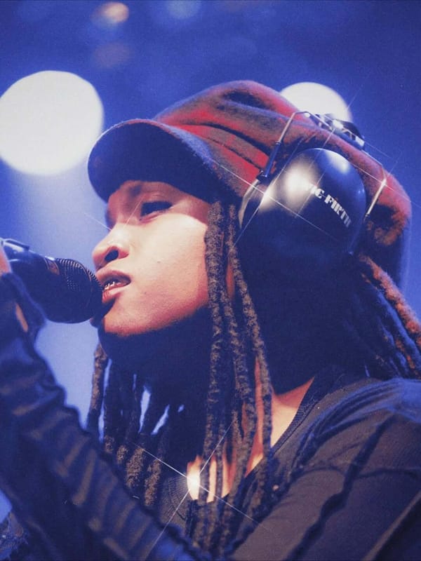 Ambré performing live on stage, singing into a microphone while wearing headphones and a knit hat, with blue and purple concert lighting. 